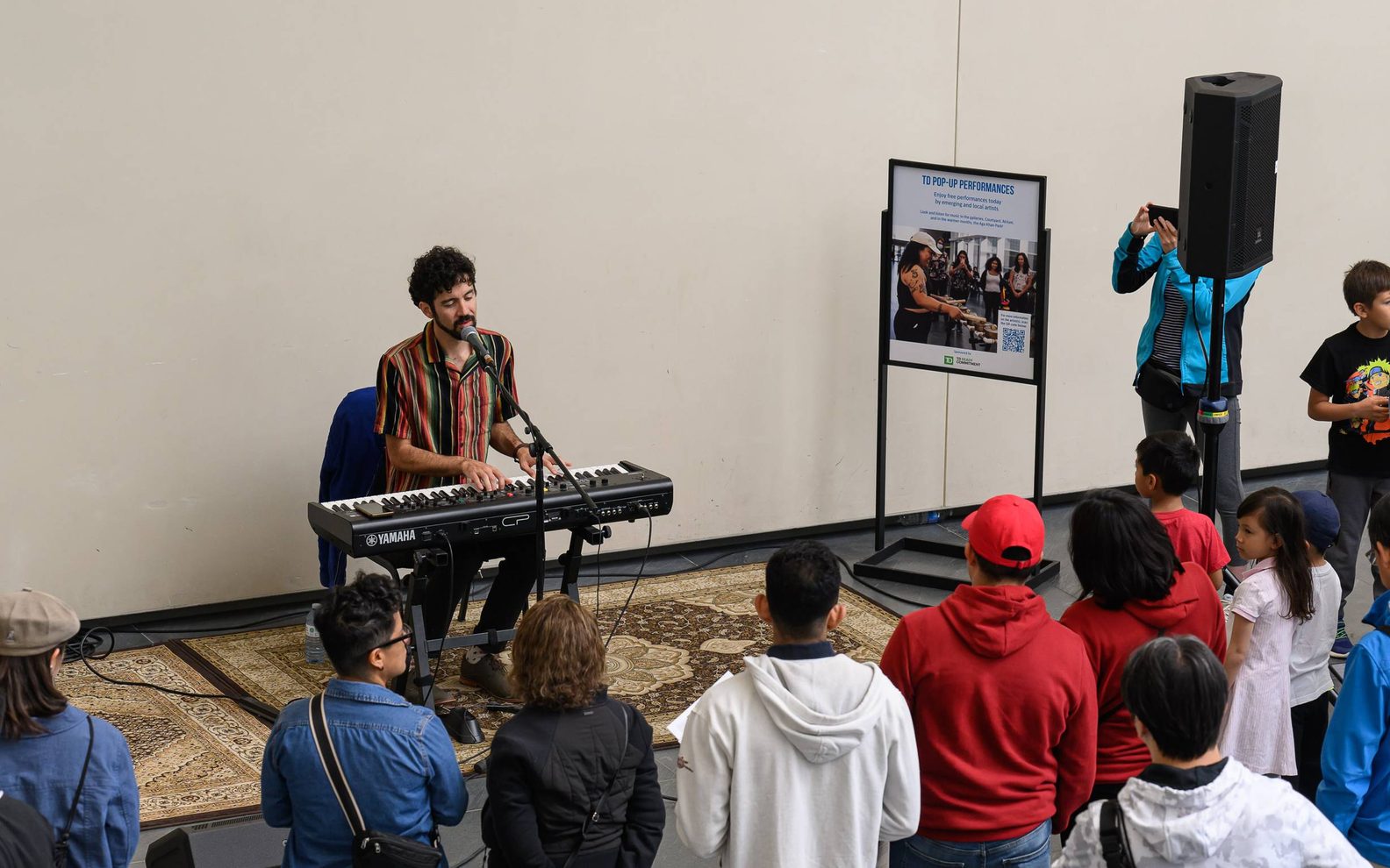 A TD pop up performer playing a piano and singing with an audience gathered in front of him inside the Aga Khan Museum