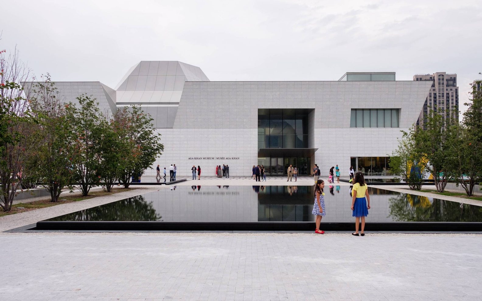 Wide exterior view of the Aga Khan Museum main entrance from over a reflecting pool on a cloudy day with children and many people gathered in front