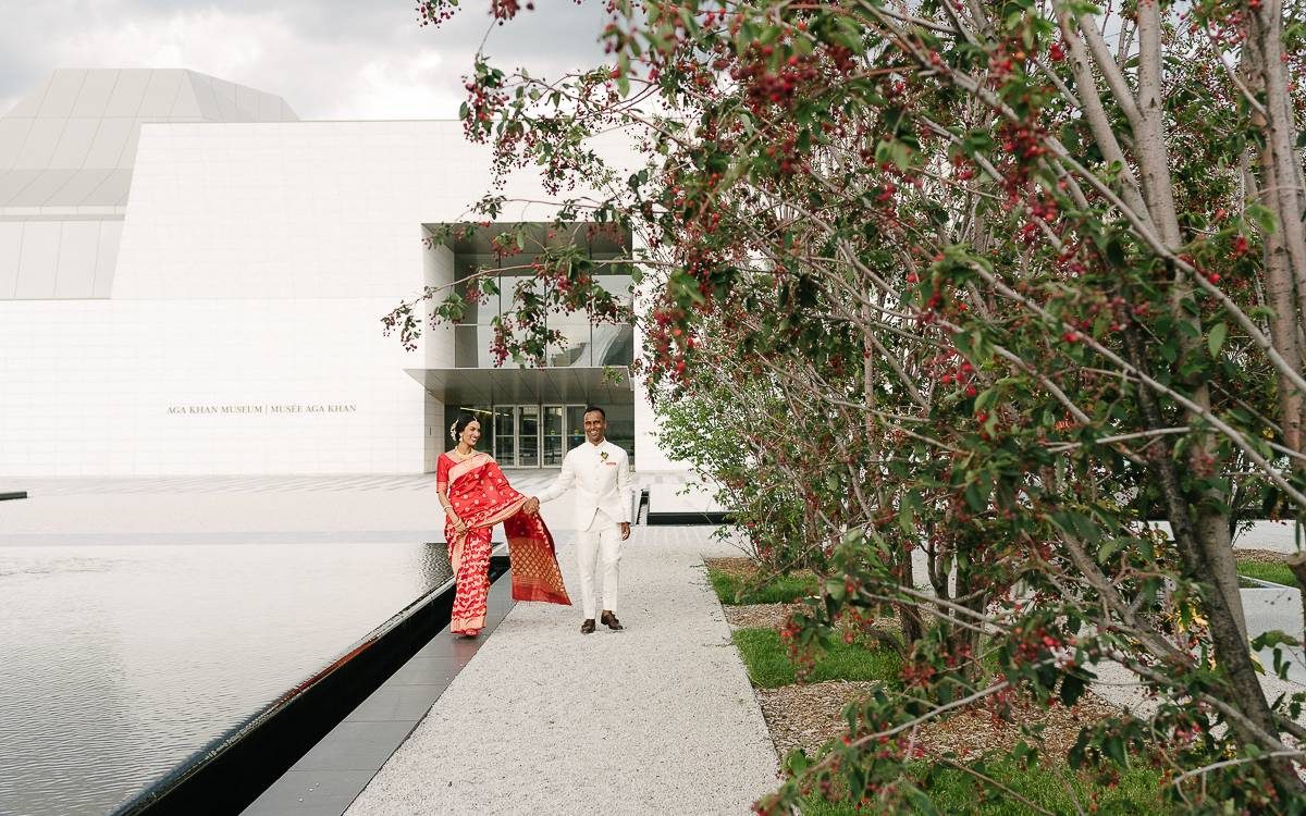Two newly weds walking by the trees in the Aga Khan Park with the front entrance to the Aga Khan Museum behind them