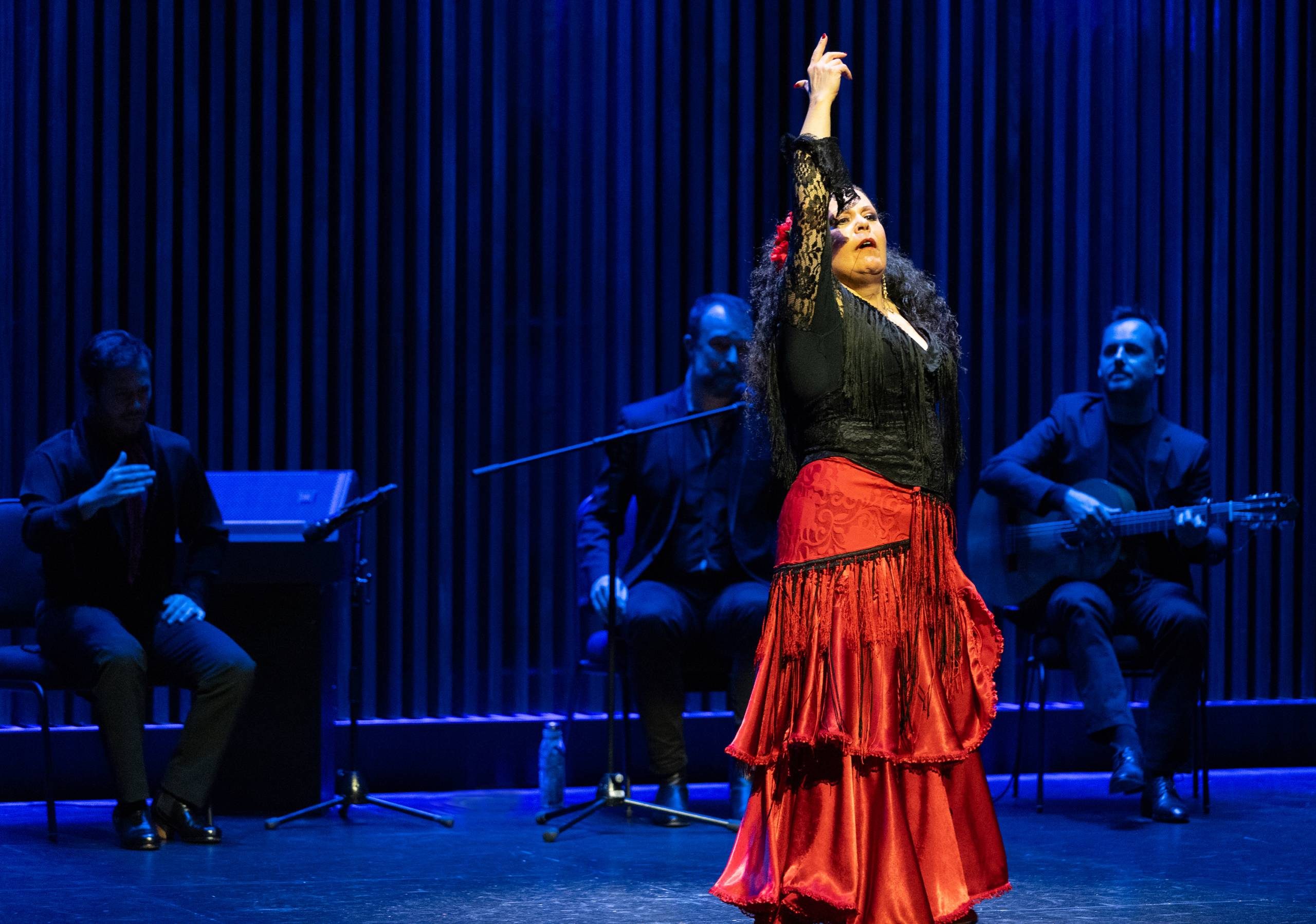 Flamenco dancer with her arm up mid dance at the Aga Khan Museum auditorium. There are musicians sitting behind her in the blue dim shadows.