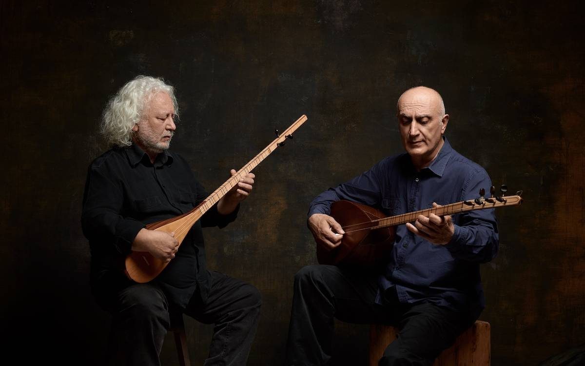 Erkan Ogur and Ismail Hakki Demircioglu playing lutes side by side on a dark gray background. Dramatic lighting and they appear to be entranced by the music