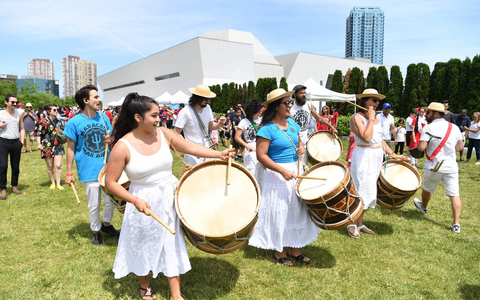 Young adults in casual white and blue clothing, marching with drums and smiling in front of the Aga Khan Museum