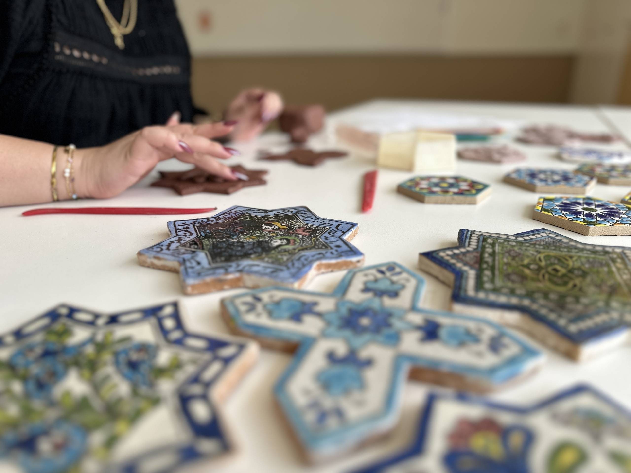 A close up of painted clay tiles with adult hands forming tiles out of clay in the back.