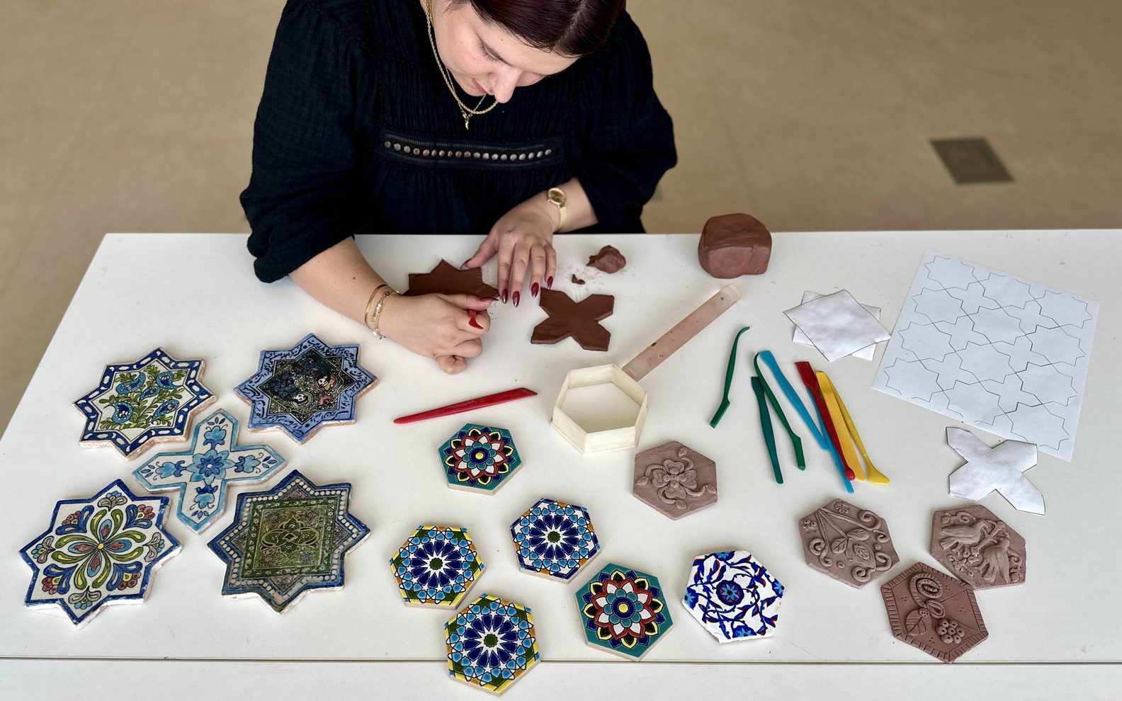 A bird's eye view of an adult shaping clay into a star-shaped tile on a table with tools and finished and decorated ceramic tiles.