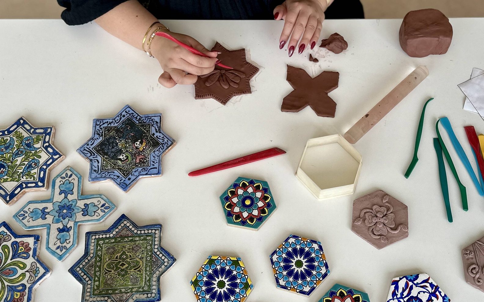 A bird's eye view of an adult imprinting patterns in clay at a table with finished, painted tiles.