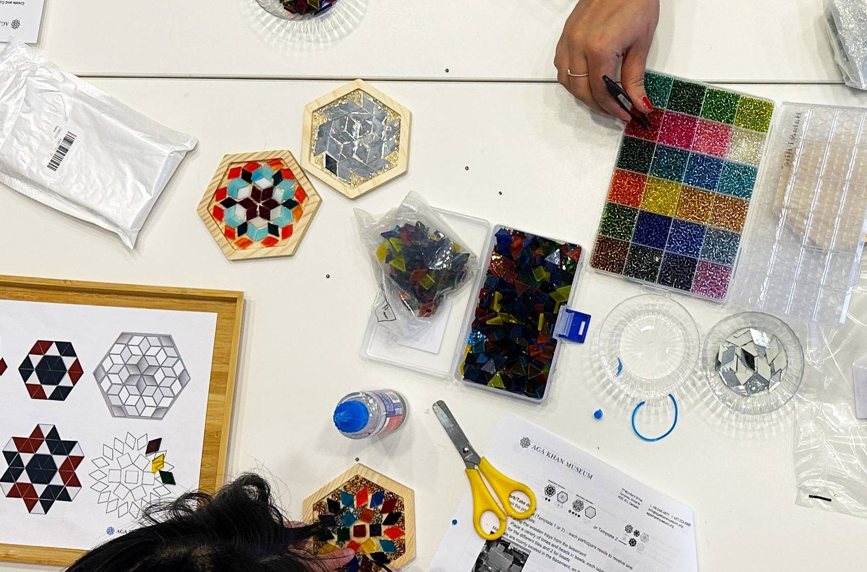 A birds-eye-view of three workshop participants creating mosaic tiles at a table with glass pieces, beads, glue, and tweezers.