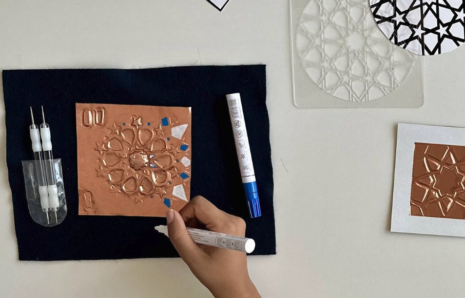 A bird's eye view of a hand painting a sheet of embossed metal among stencils, patterned paper, and embossing tools.