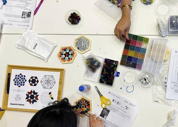 A bird's eye view of a table with three adults creating mosaic tiles out of glass pieces, beads, tweezers, and glue.