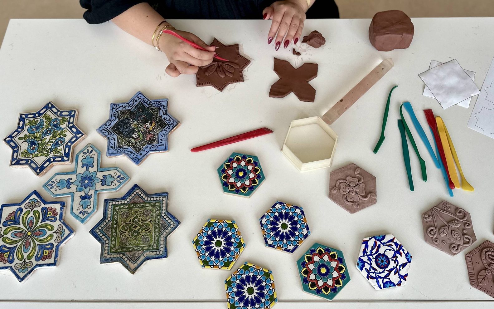 A bird's eye view of an adult imprinting patterns in clay at a table with finished, painted tiles.