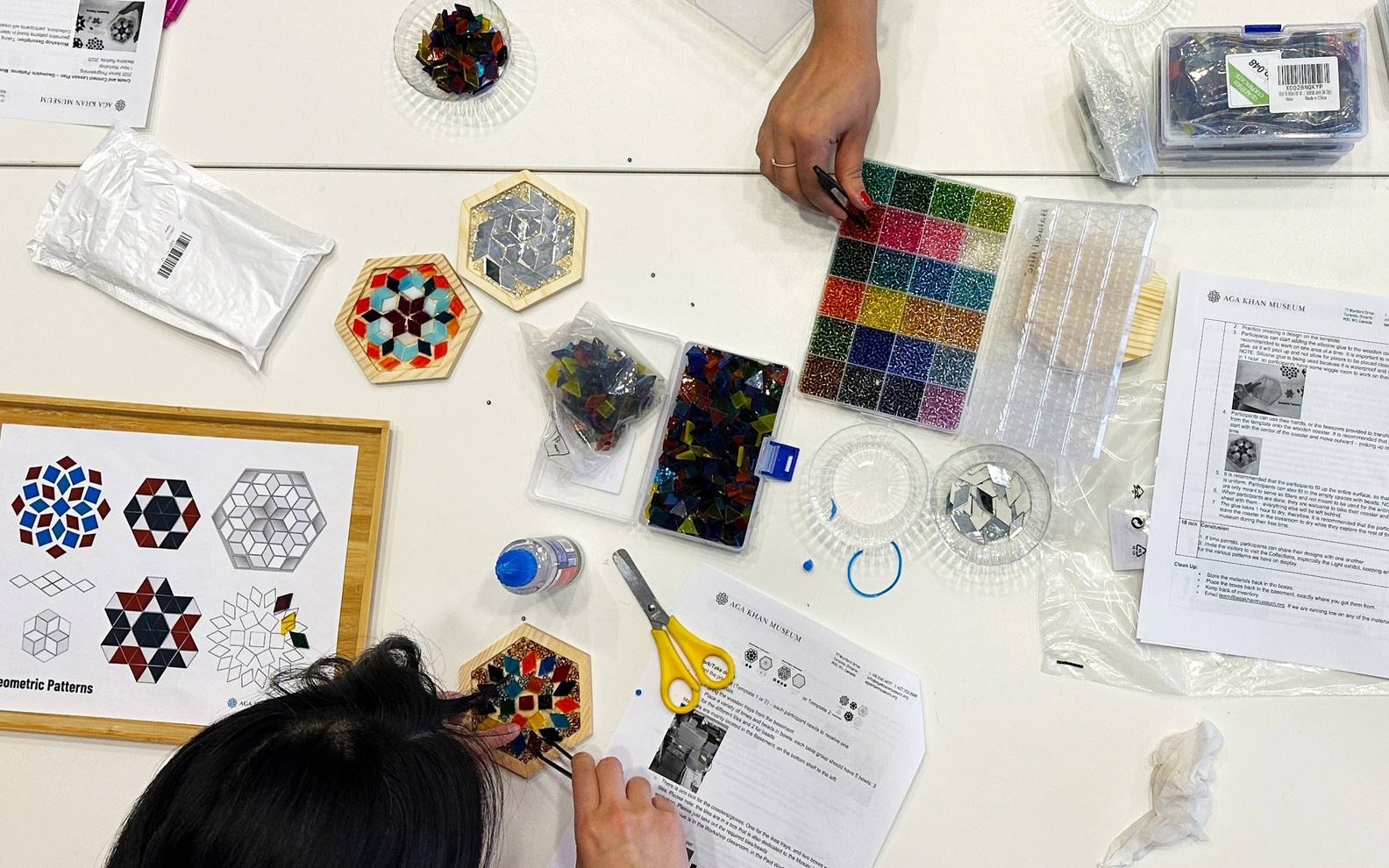 A birds-eye-view of three workshop participants creating mosaic tiles at a table with glass pieces, beads, glue, and tweezers.