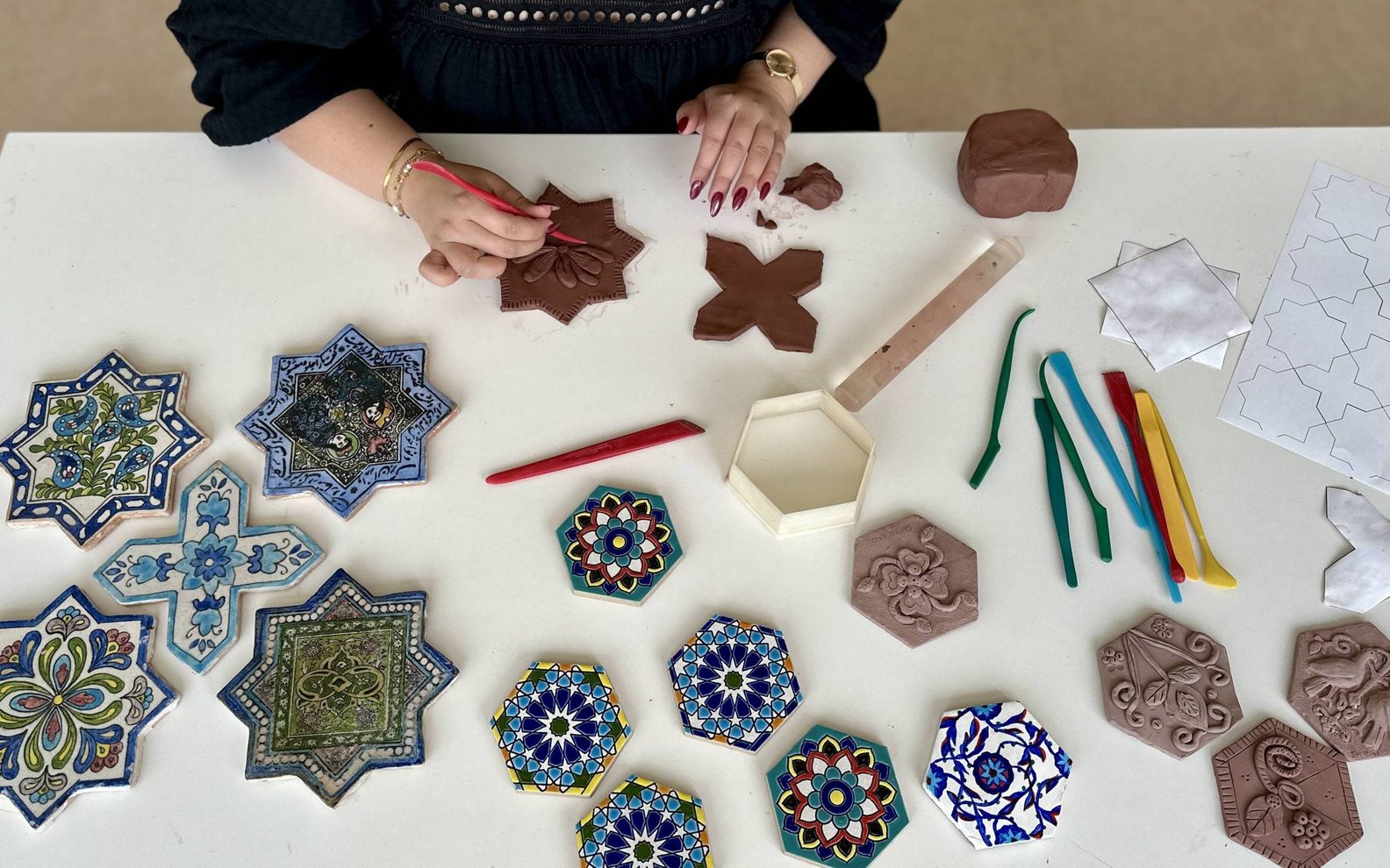 A bird's eye view of an adult imprinting patterns in clay at a table with finished, painted tiles.