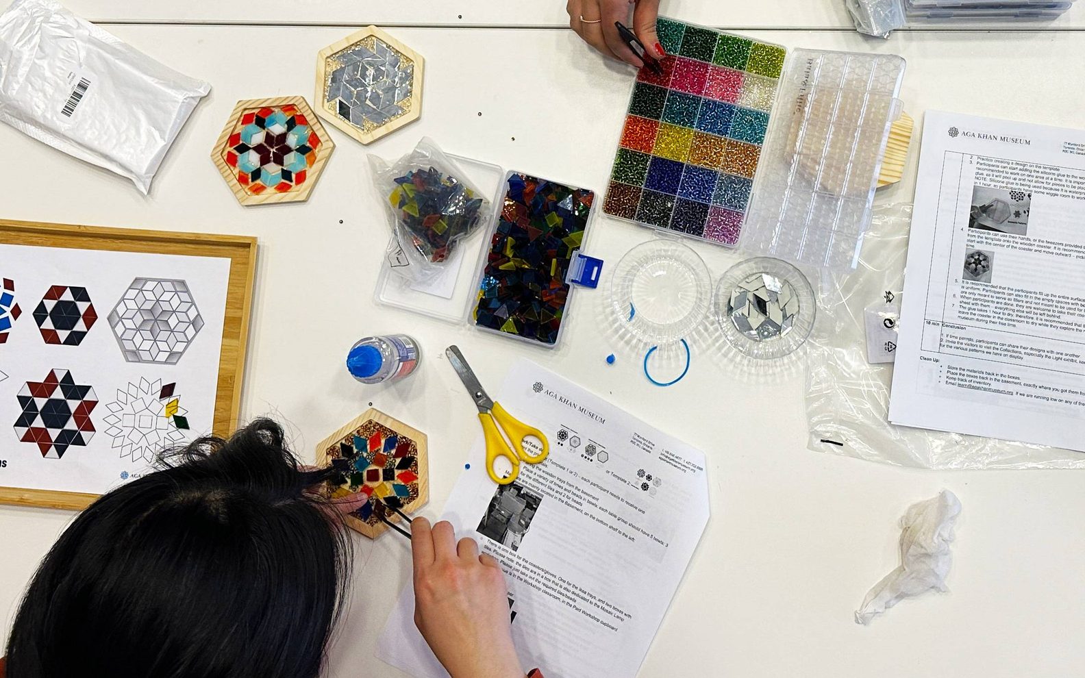 A birds-eye-view of three workshop participants creating mosaic tiles at a table with glass pieces, beads, glue, and tweezers.