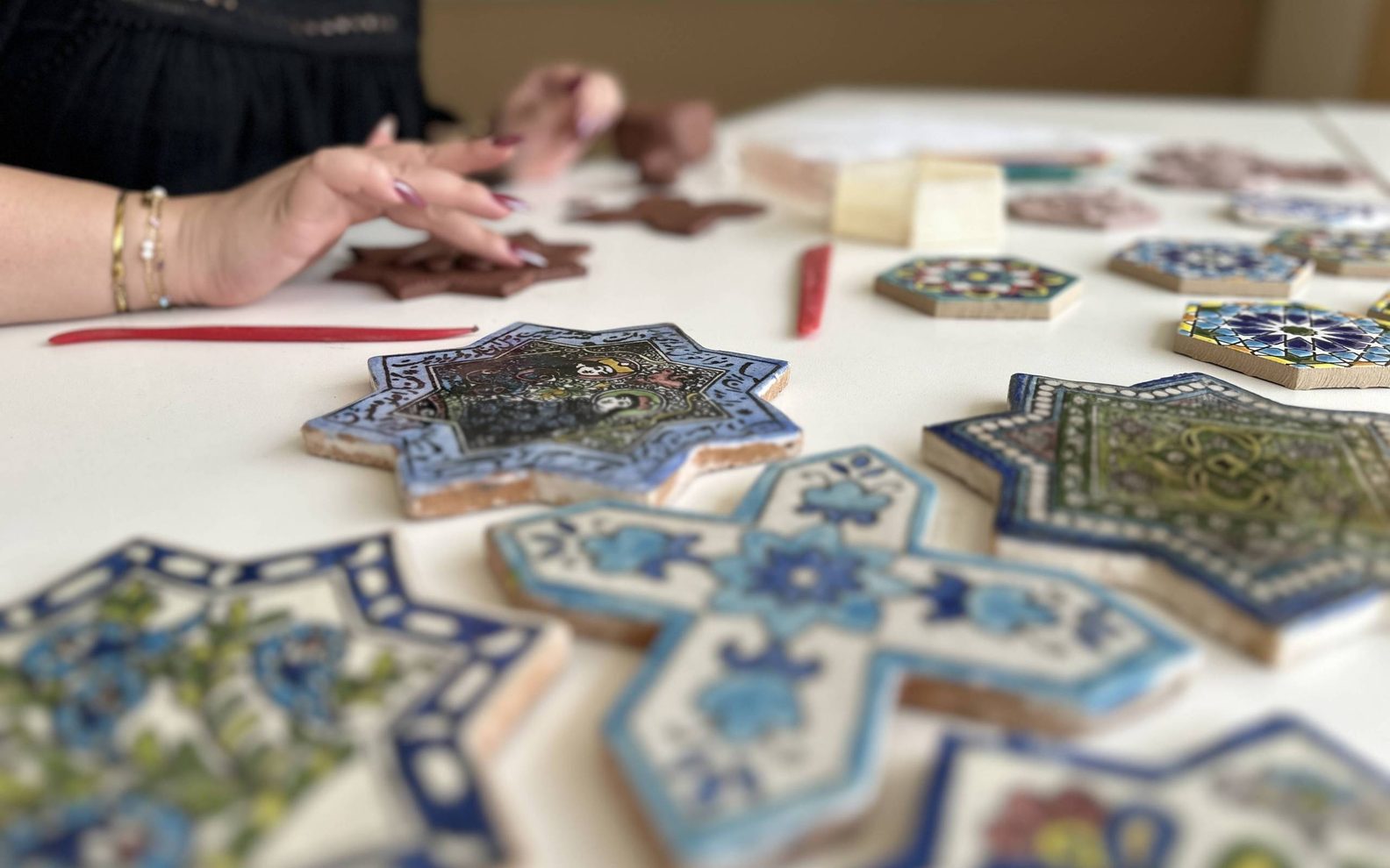 A close up of painted clay tiles with adult hands forming tiles out of clay in the back.