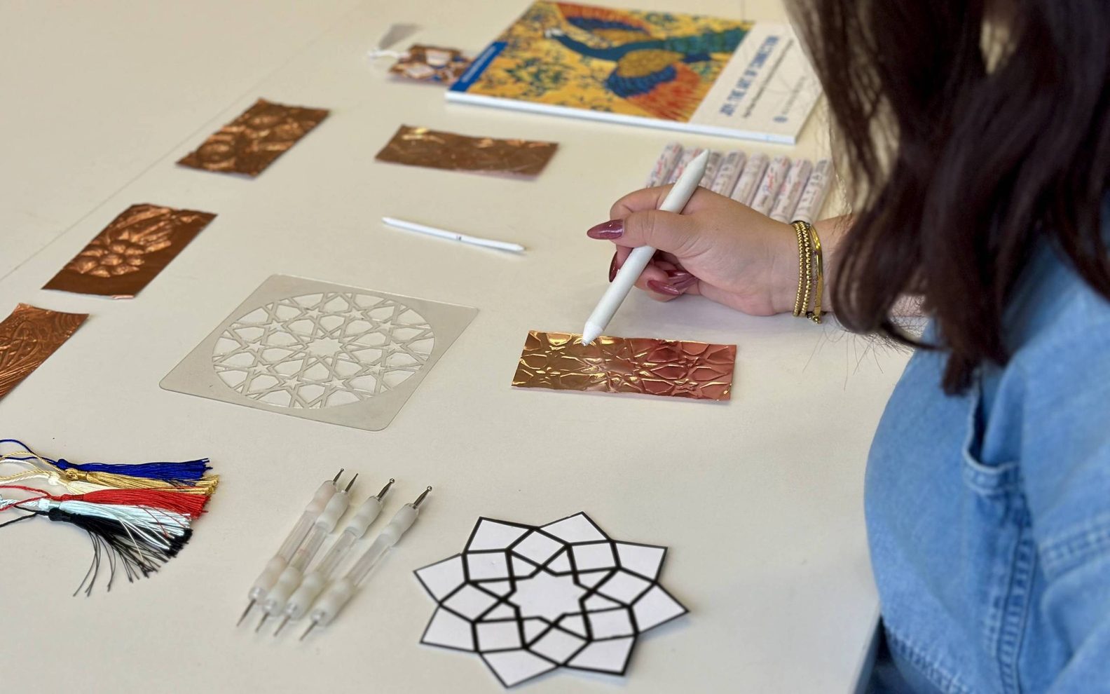 An adult sitting at a table, using tools to emboss a metallic bookmark.