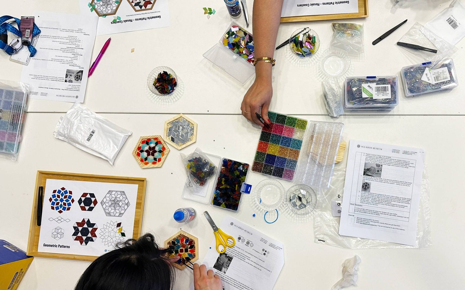 A birds-eye-view of three workshop participants creating mosaic tiles at a table with glass pieces, beads, glue, and tweezers.