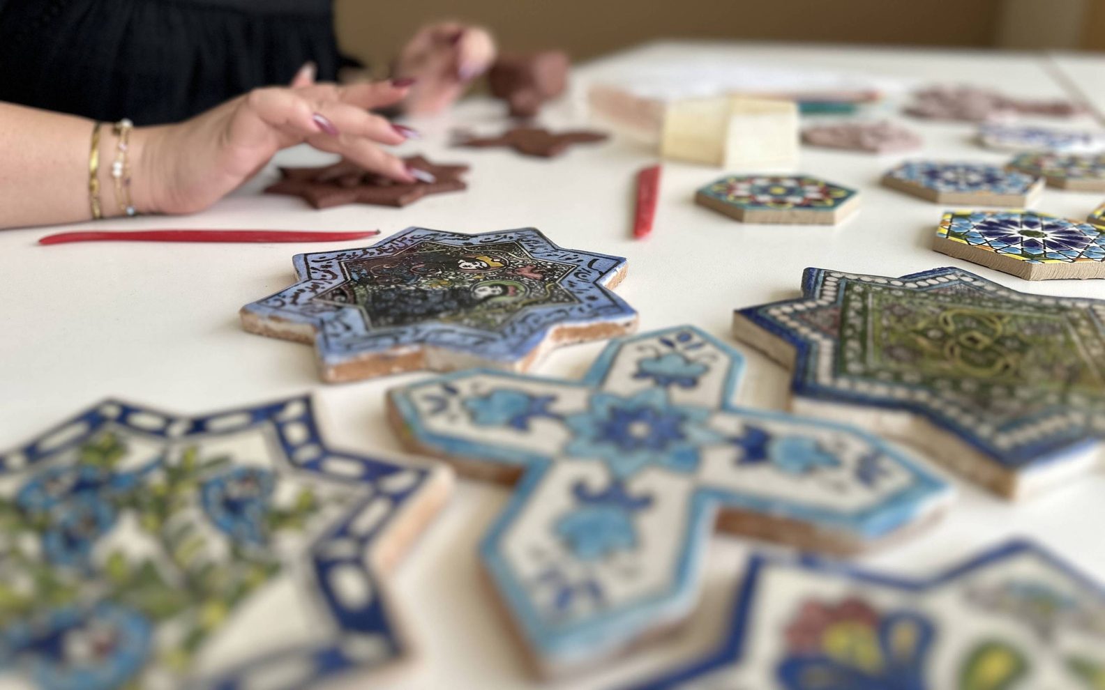 A close up of painted clay tiles with adult hands forming tiles out of clay in the back.