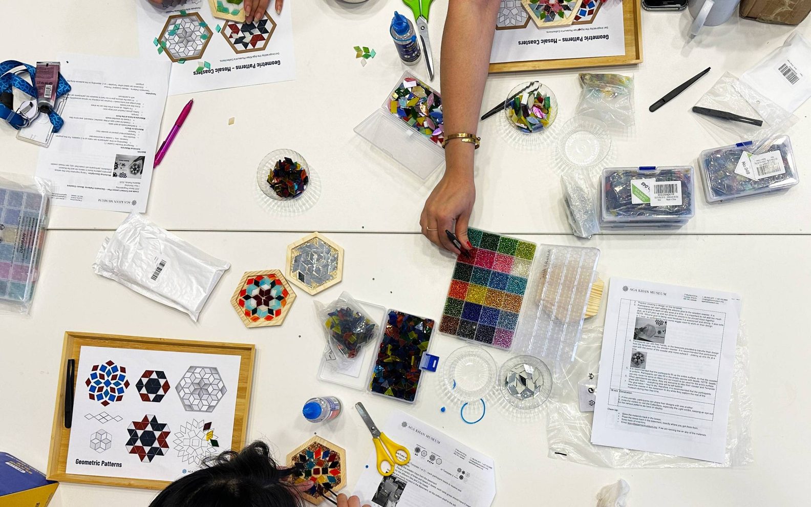 A birds-eye-view of three workshop participants creating mosaic tiles at a table with glass pieces, beads, glue, and tweezers.